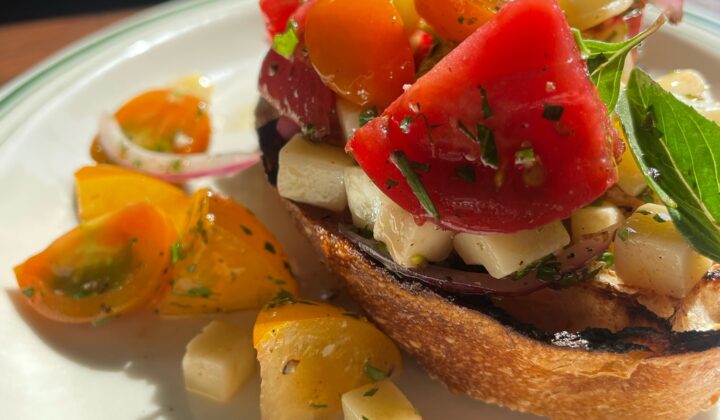 A close-up of a vibrant bruschetta dish featuring toasted bread topped with a colorful mix of chopped tomatoes, mozzarella cubes, red onions, and fresh basil. The sunny light accentuates the freshness of the ingredients. Scattered tomato pieces and herbs garnish the plate. - Hausion