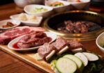 An array of raw meats and vegetables are displayed on a wooden board beside a tabletop grill. Bowls of condiments and sides surround the setup. Meat cuts include marbled beef and ribs, while vegetables include zucchini and mushrooms. The grill has cooking meat on it. - Hausion