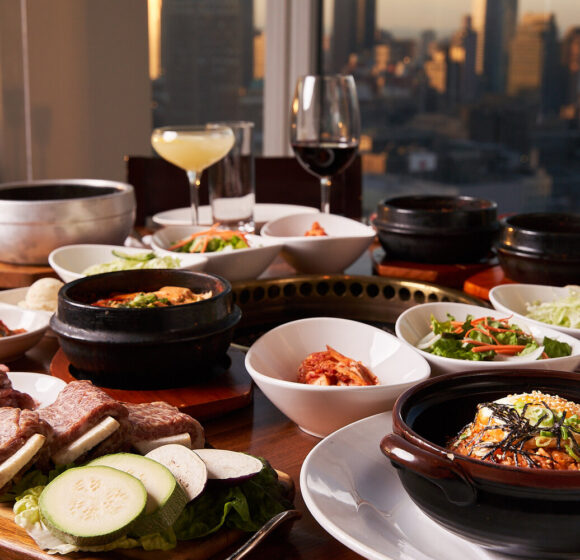 A table set with various Korean dishes including kimchi, vegetables, soups, and grilled meats, against the backdrop of a city skyline. Wine glasses and a cocktail are also present on the table. - Hausion