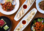 An assortment of dishes on a wooden table. The centerpiece consists of dumplings served with soy sauce in white elongated plates. Surrounding dishes include a variety of meats, vegetables, and a salad, presenting a diverse and colorful meal spread. - Hausion