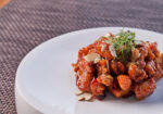 A dish of caramelized honey chicken garnished with sliced almonds and herbs is served on a white plate. The textured, brown placemat beneath the plate adds a subtle background contrast. - Hausion