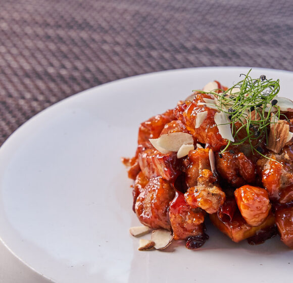 A dish of caramelized honey chicken garnished with sliced almonds and herbs is served on a white plate. The textured, brown placemat beneath the plate adds a subtle background contrast. - Hausion