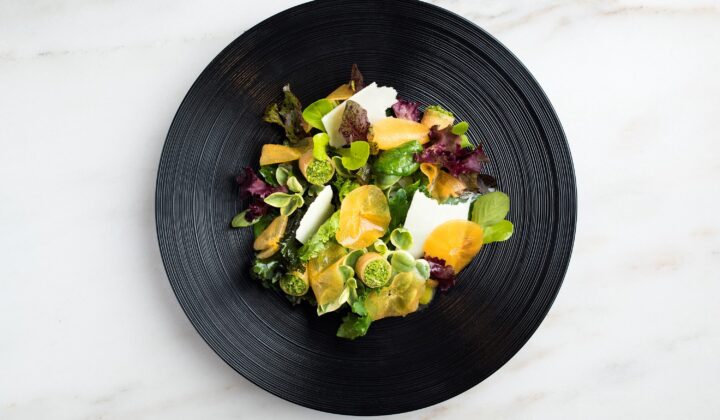 A gourmet salad served on a black plate, featuring a vibrant mix of leafy greens, thinly sliced citrus fruits, possibly oranges, and garnished with small round green elements. The dish is aesthetically presented against a marble surface. - Hausion